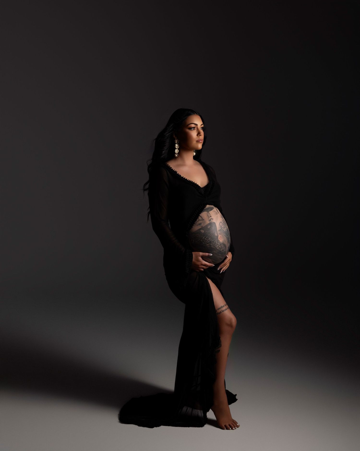 Artistic maternity photo of an expectant mother in a flowing black dress against a dark studio backdrop.