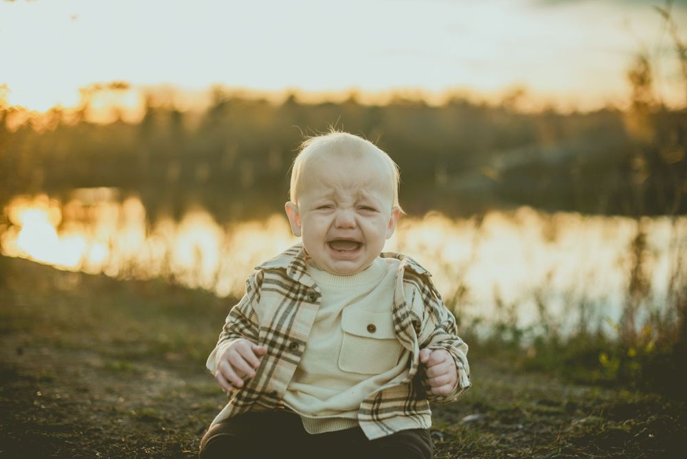 Baby wearing plaid flannel jacket laughs joyfully by a lake at sunset.