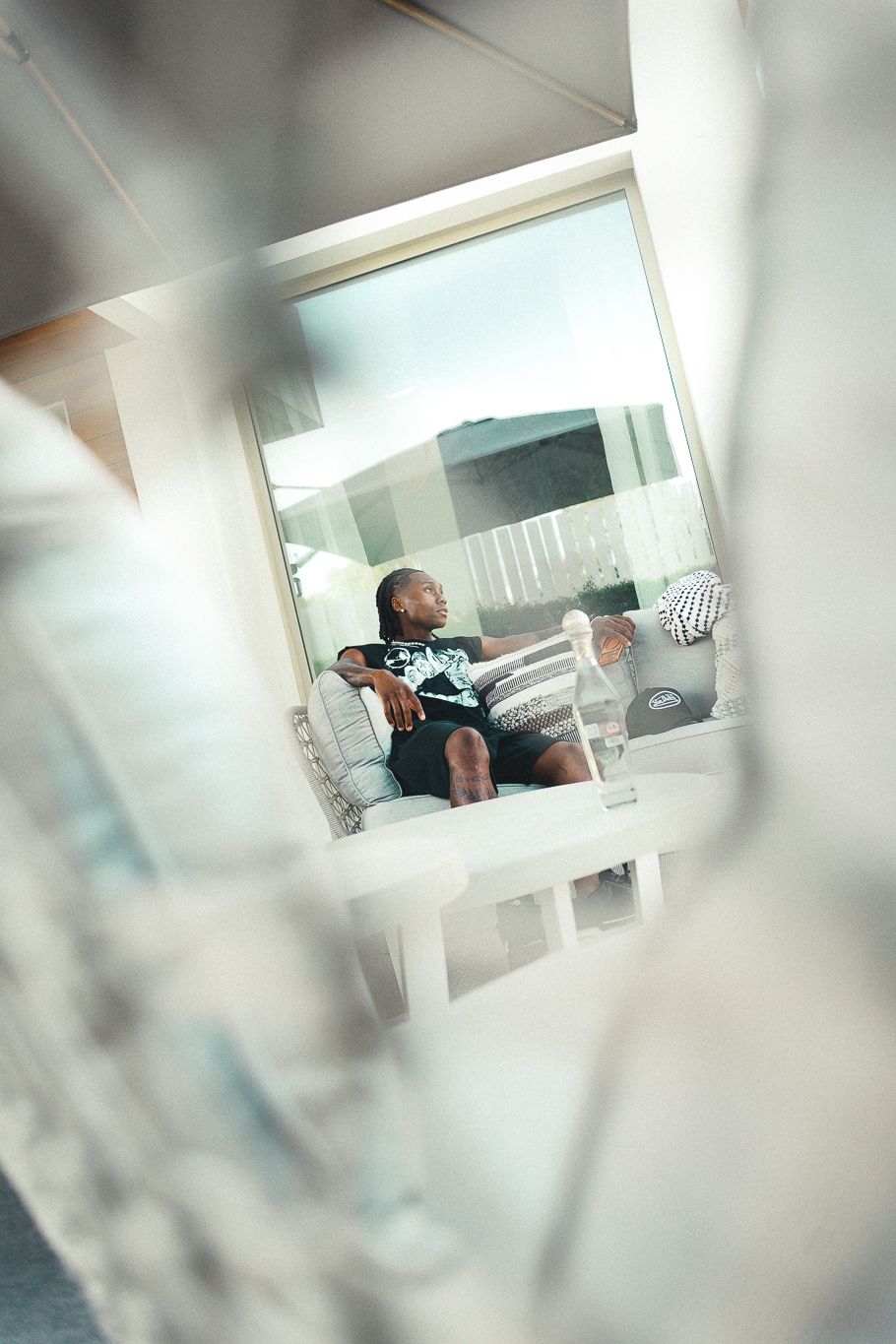 Man relaxing on a white sofa near a large window, viewed through a blurred foreground in a bright modern interior.