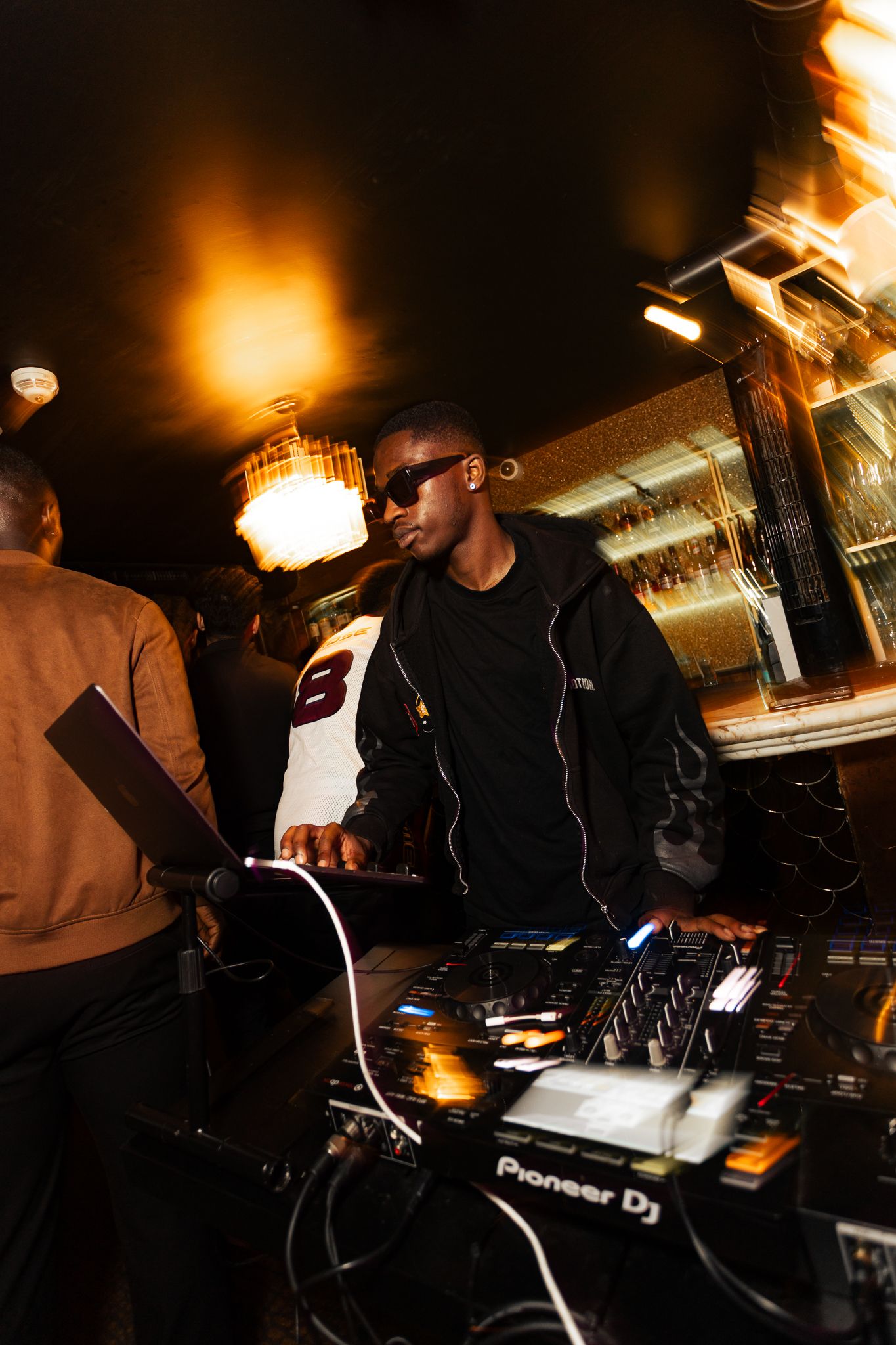 DJ performs at a dimly lit nightclub using Pioneer DJ equipment while patrons watch in the background.