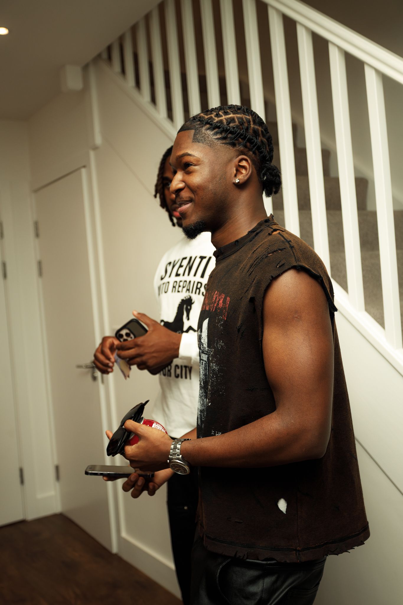 Person in a brown sleeveless shirt smiling while standing near white stairs indoors.