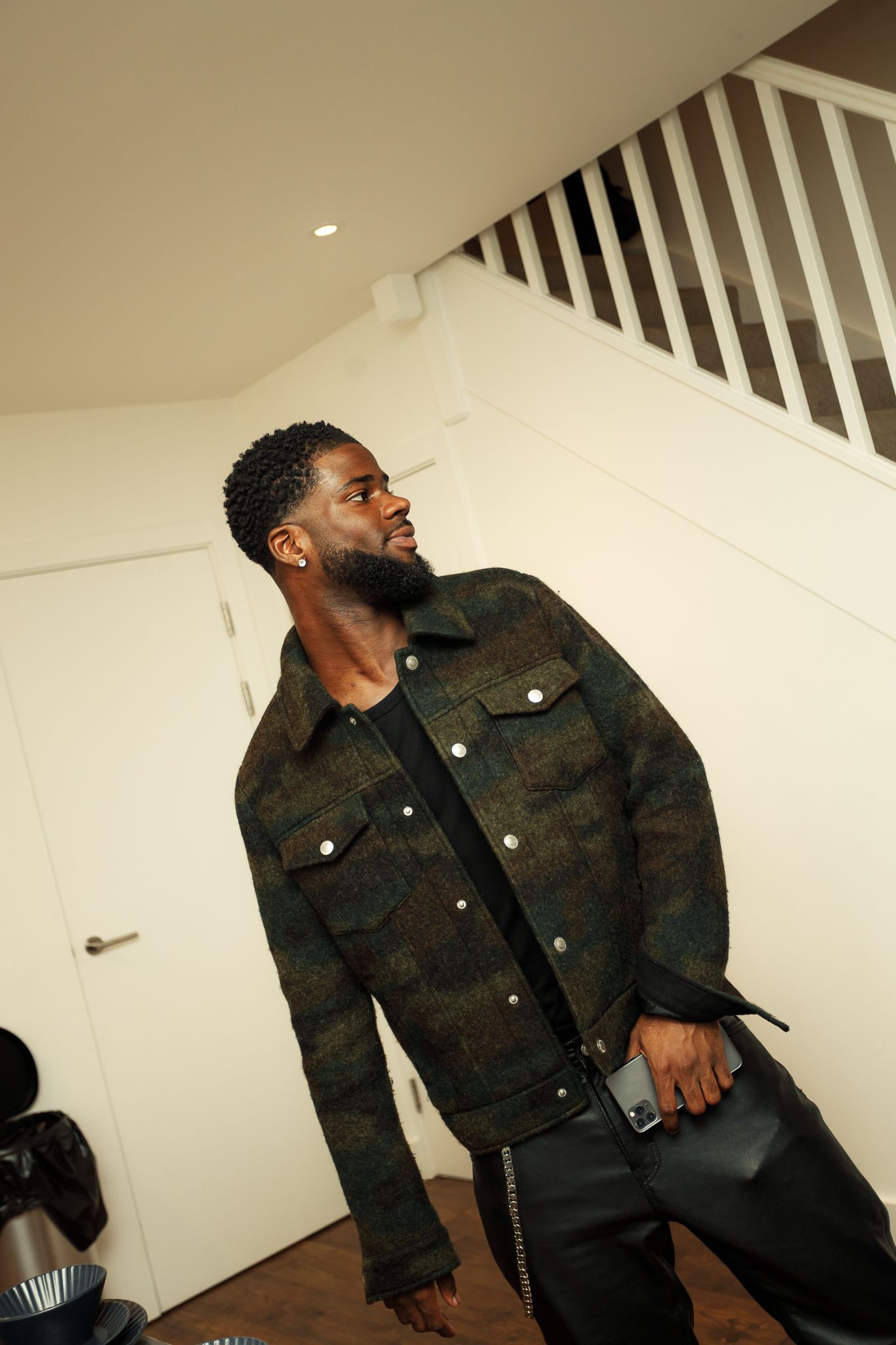 A person wearing a dark camouflage jacket and black pants poses against a white wall with stairs in the background.