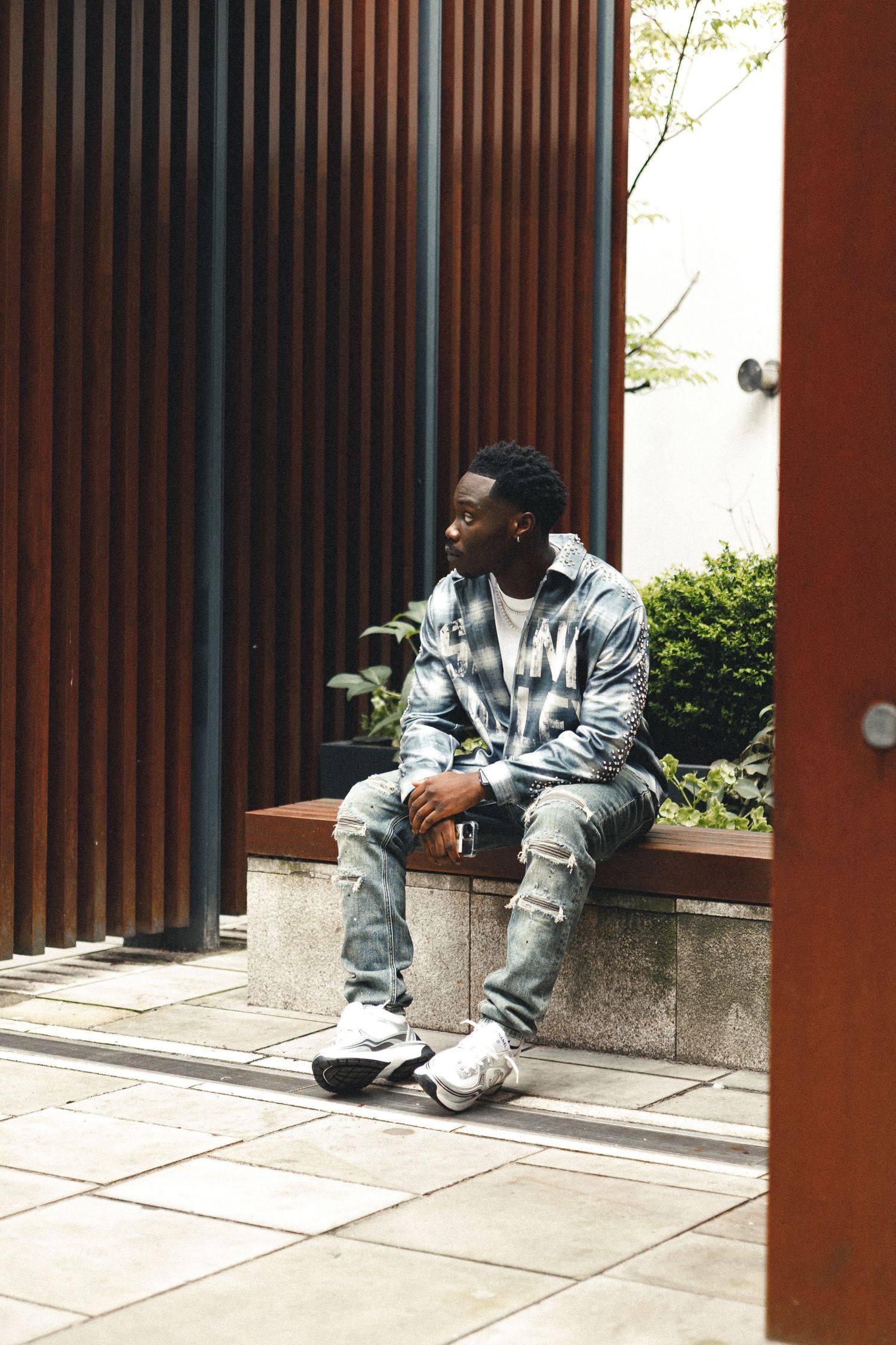 A young Black man sits on a bench outdoors, wearing a plaid shirt and jeans, holding a drink, with wooden panels behind him.