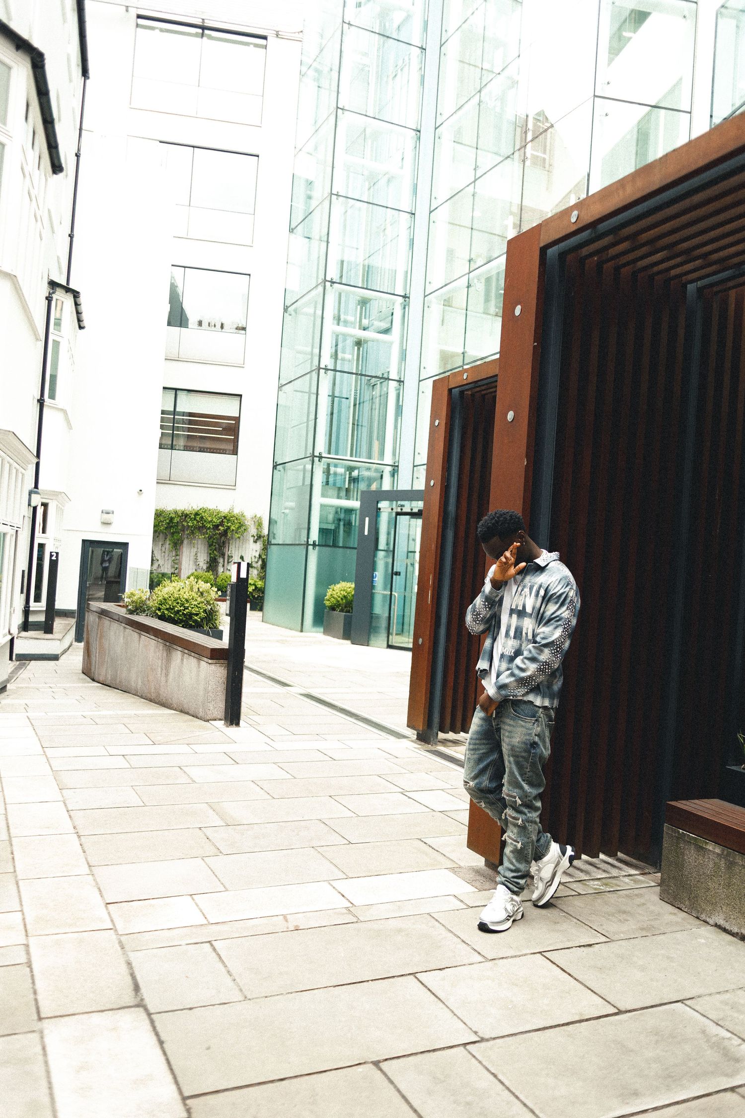 A young man in casual streetwear leans against a modern glass building on a paved urban alley, looking at his phone.