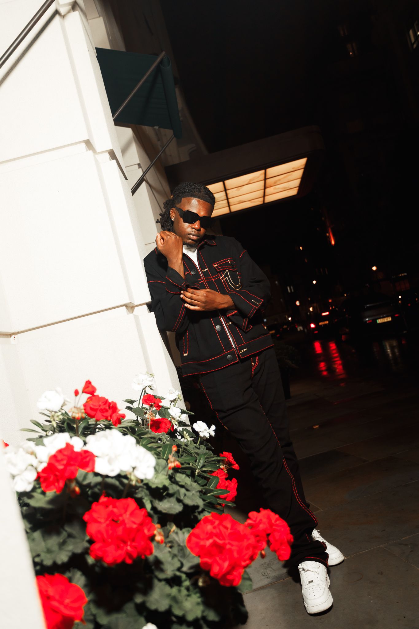 Person in black outfit and sunglasses poses near red and white flowers at night.