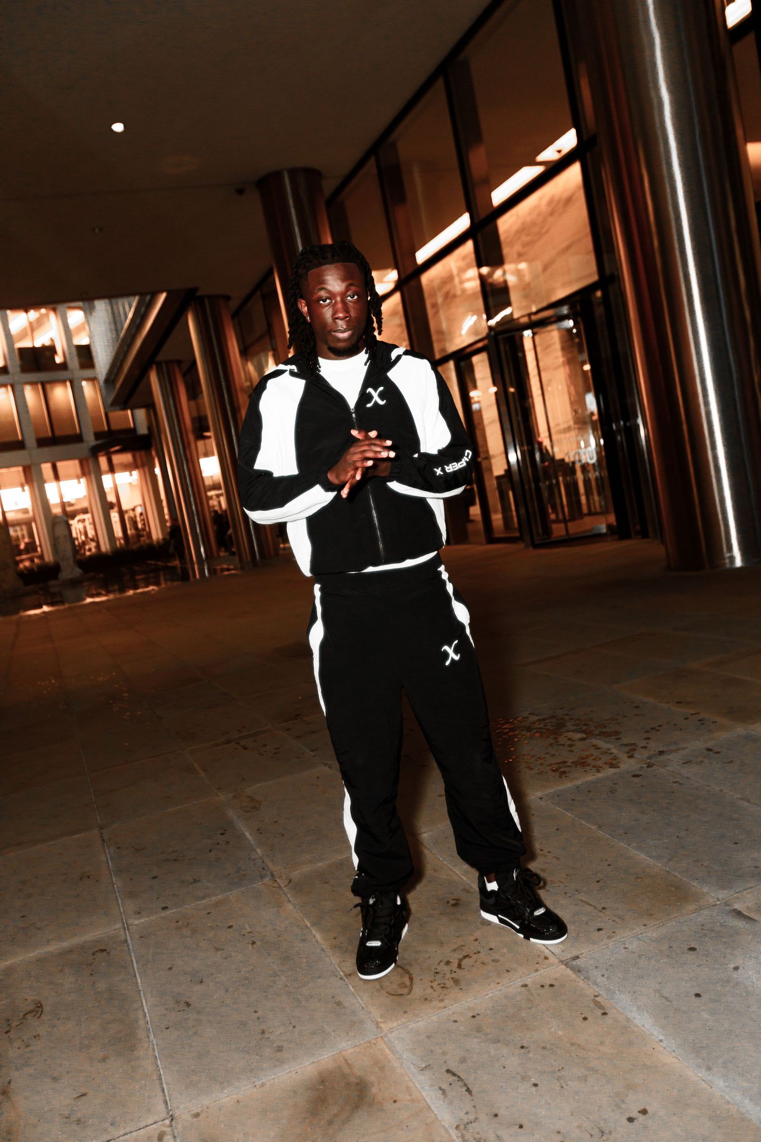 A person wearing black and white clothing stands in a dimly lit corridor with columns and ambient lighting.