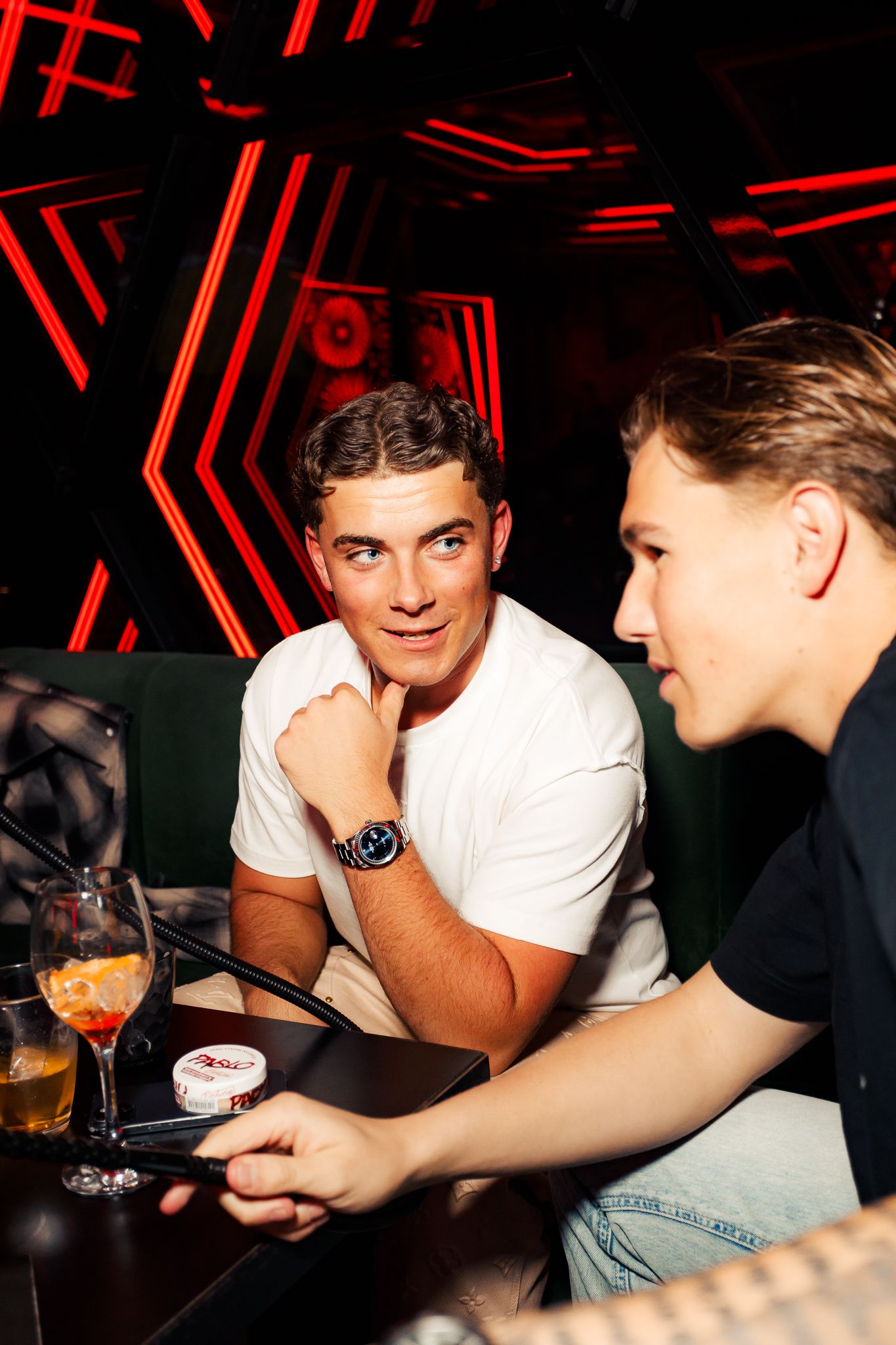 Two people having drinks at a dimly lit bar with red neon lighting in the background.