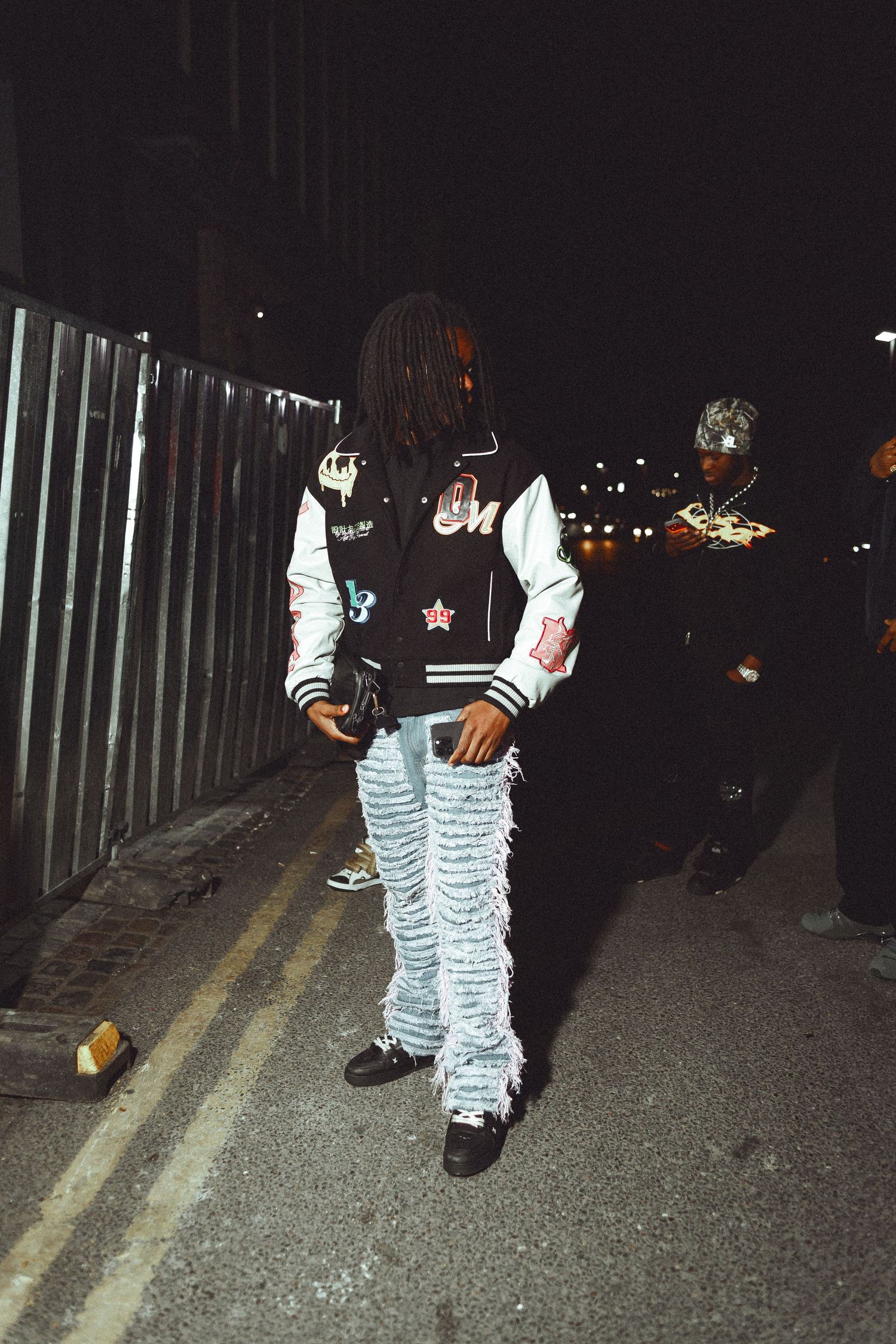 Person wearing a varsity jacket with patches and distressed fringe jeans stands on a sidewalk at night.