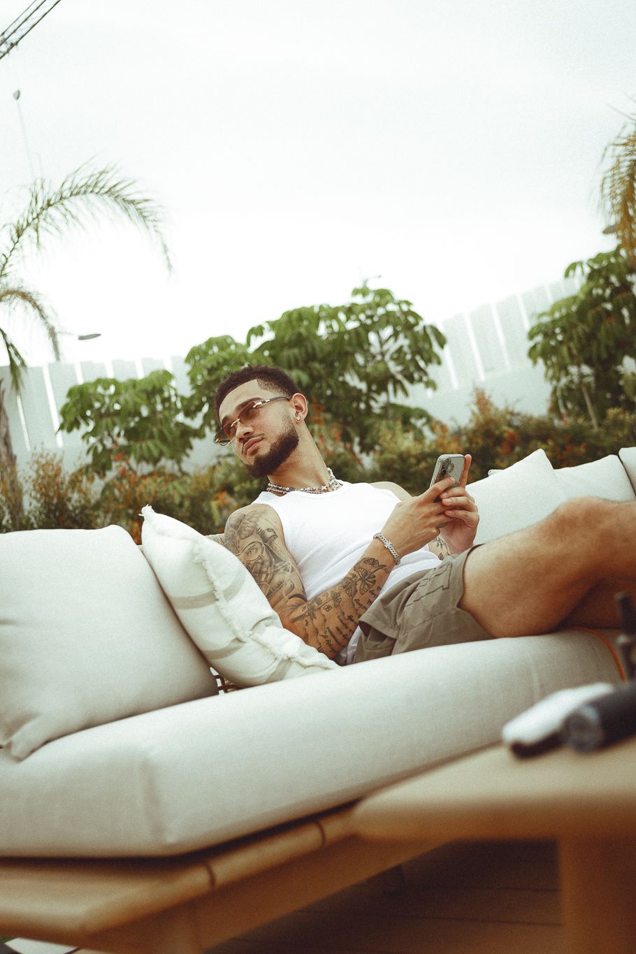 Tattooed man in sunglasses relaxing on outdoor sofa, using smartphone, with tropical greenery in background.
