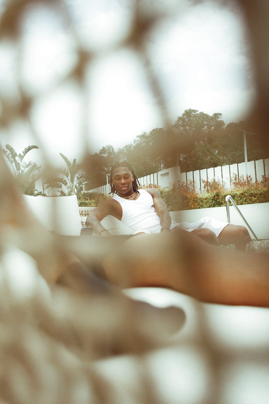 Man relaxing poolside in white tank top, viewed through a net on a sunny day.
