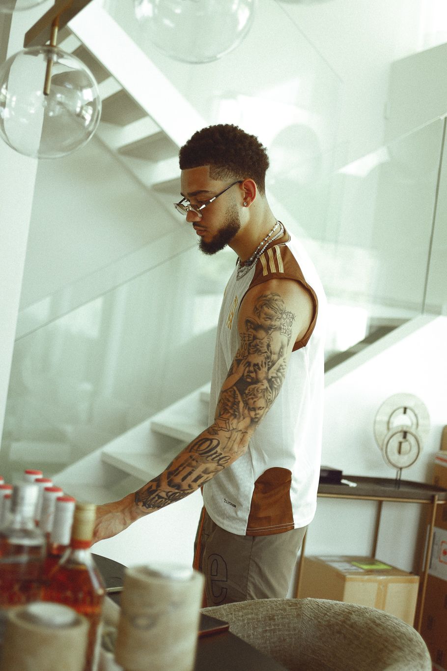 Tattooed man in white sleeveless shirt and glasses standing at a kitchen counter in a modern home.