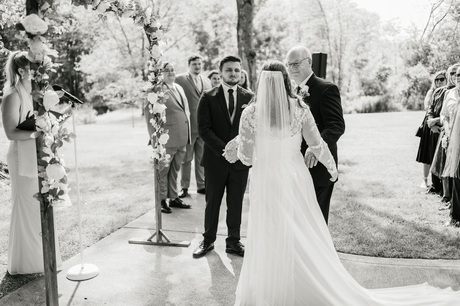 Black and white outdoor wedding ceremony with a bride in a flowing veil standing at the altar surrounded by guests.