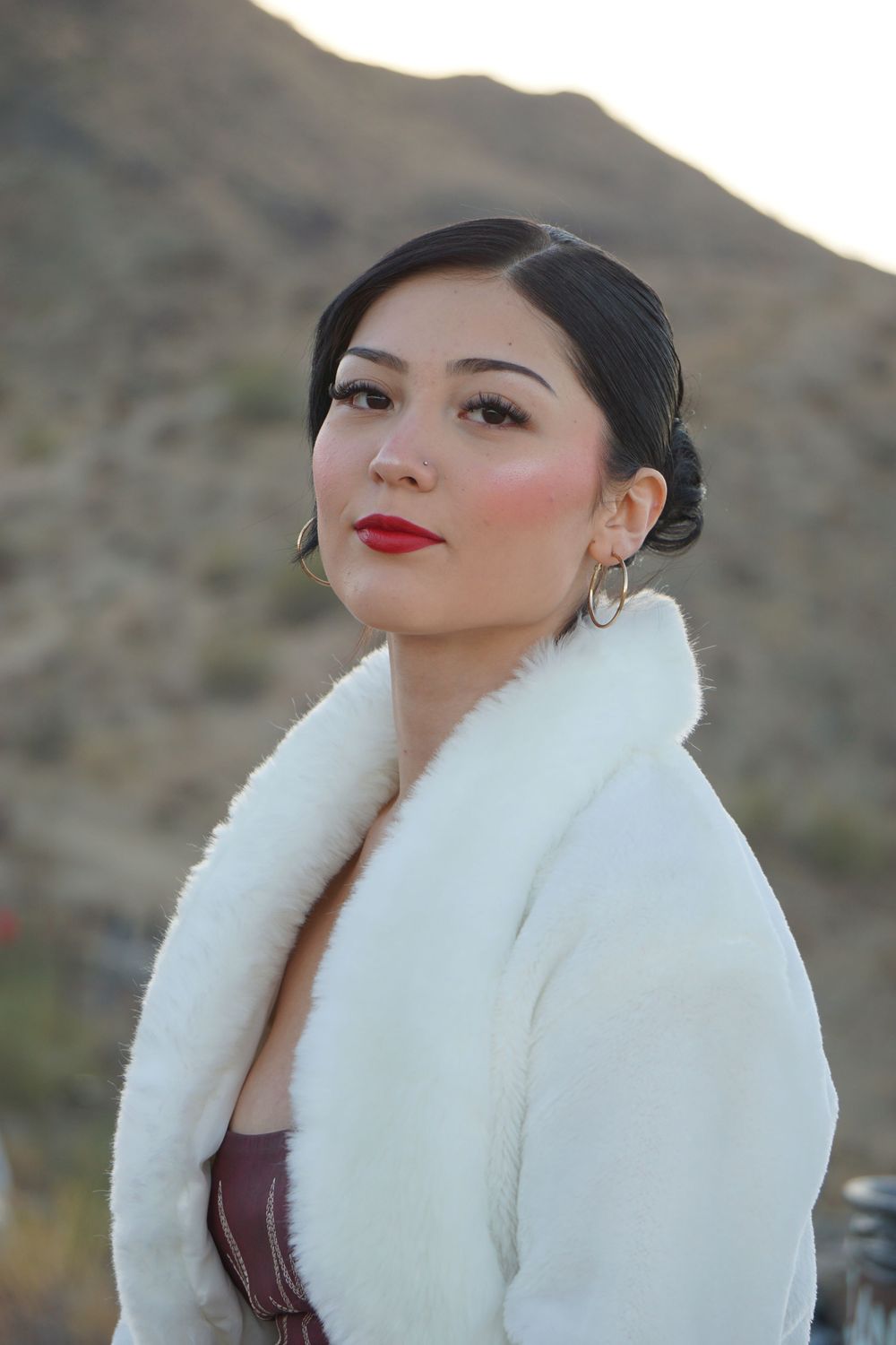 A woman in a white fur collar coat poses elegantly against a desert mountain backdrop.