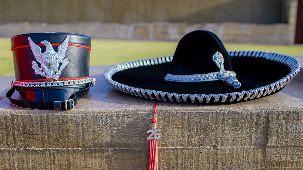 A military shako hat and black mariachi sombrero displayed side by side on a stone ledge.