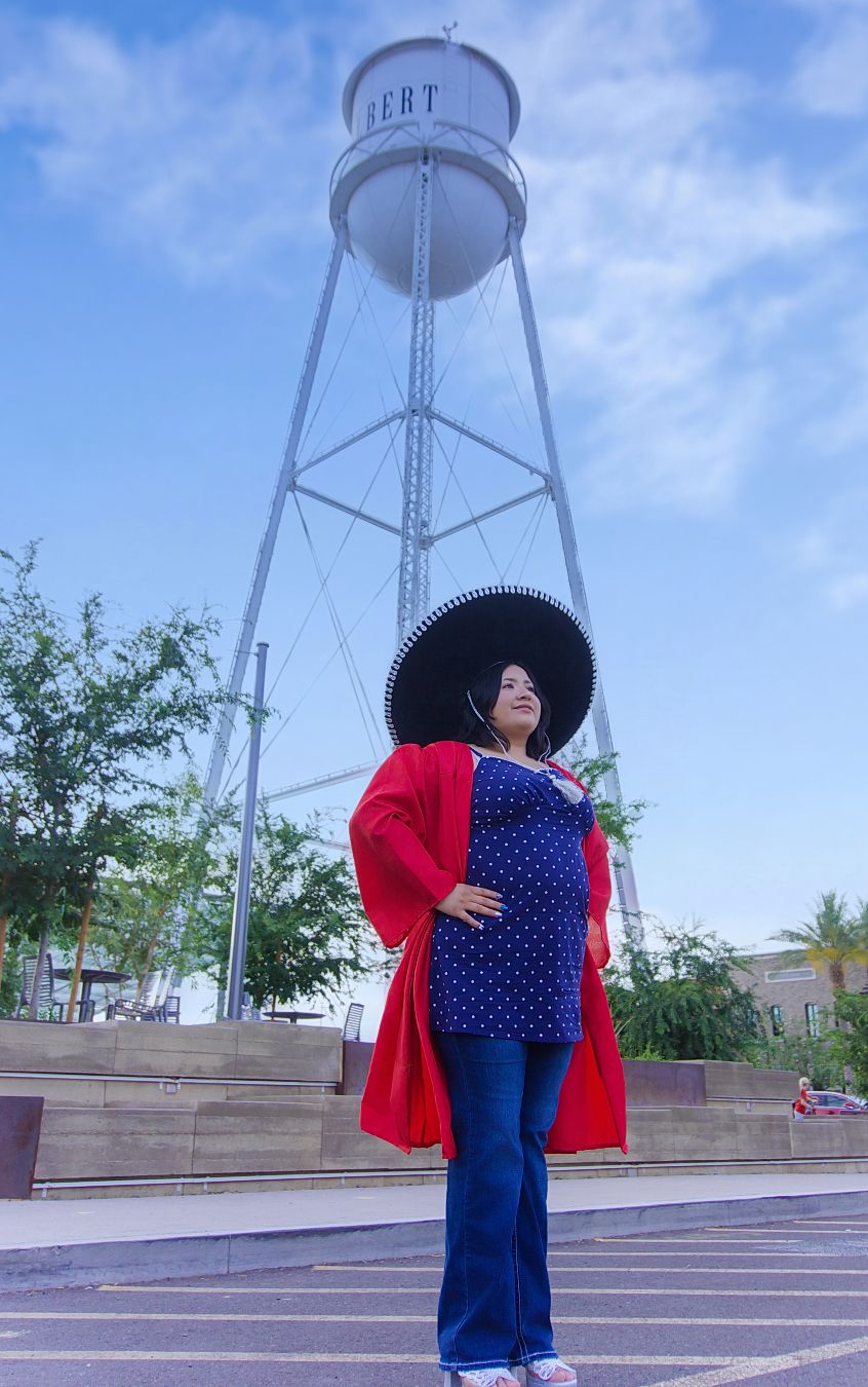 Pregnant woman in red jacket, blue top, jeans, and sombrero poses before a water tower on a sunny day.