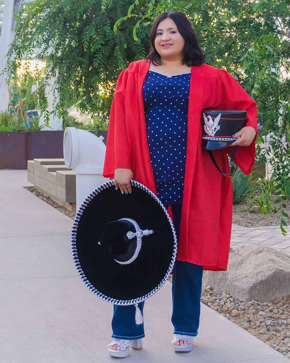 Woman in red graduation gown holds black sombrero and purse, wearing blue polka dot dress and jeans outdoors.