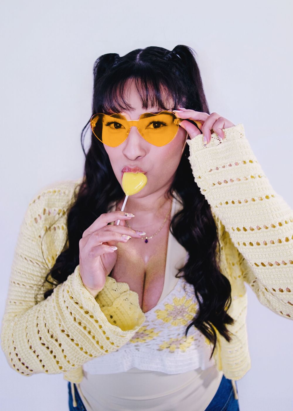 A person in yellow tinted glasses and cream knit cardigan poses playfully with a lollipop against white background.