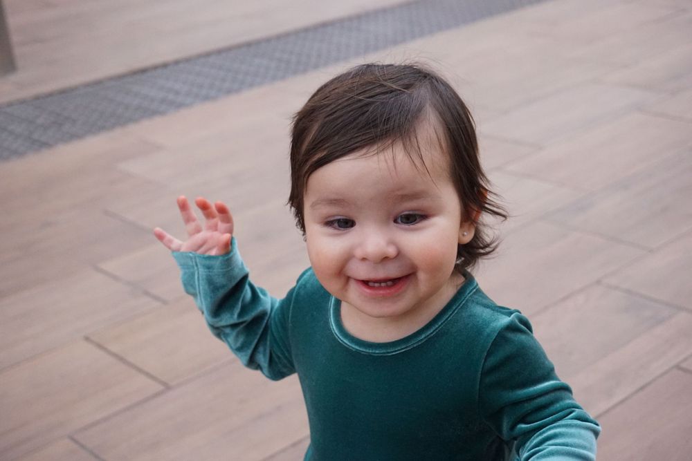 Young child in teal dress waving and smiling on hardwood floor.