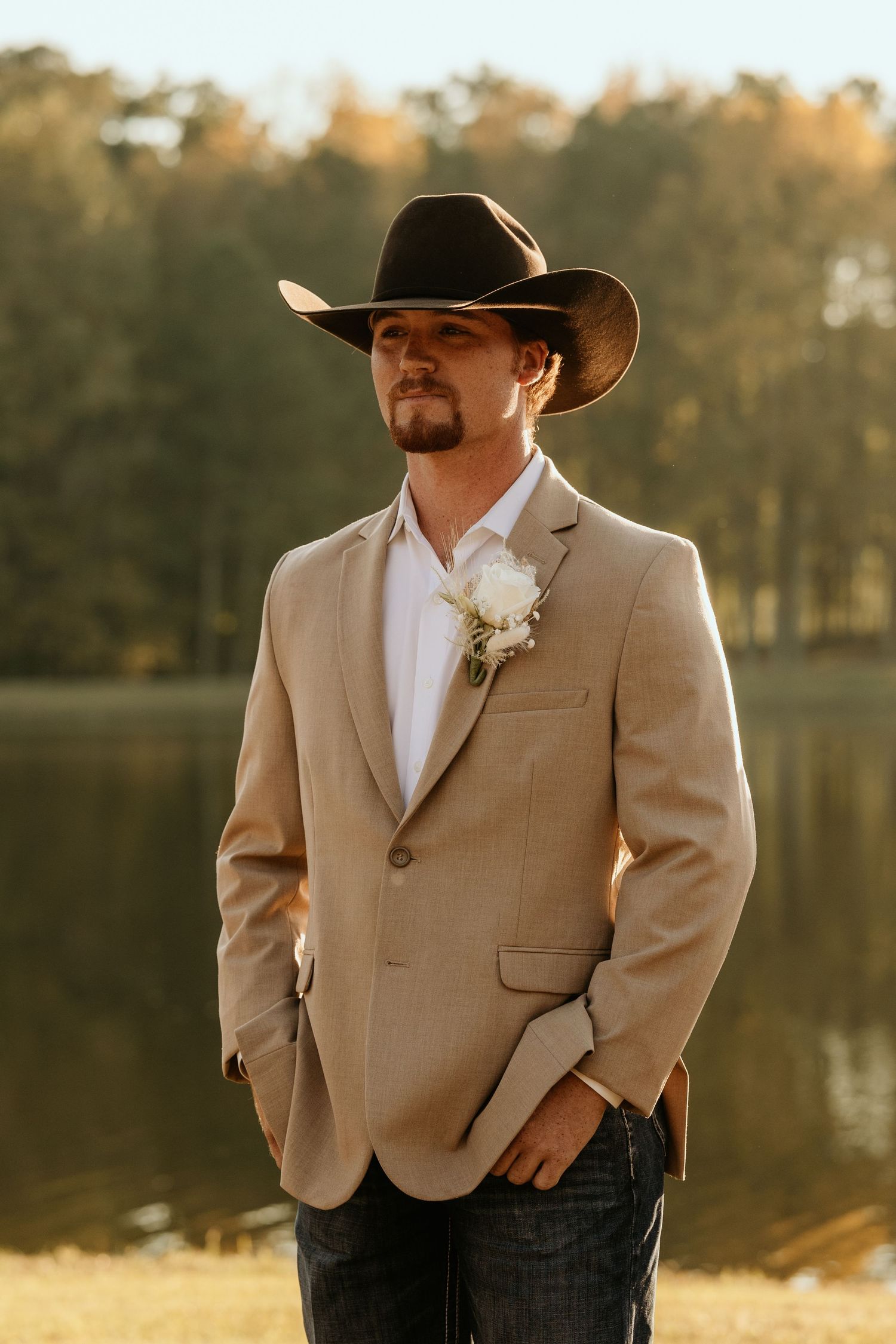 Groom in tan blazer and cowboy hat stands by lake during golden hour.