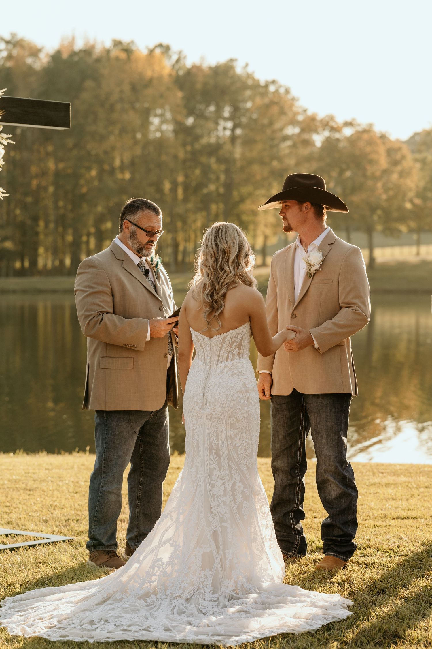Outdoor lakeside wedding ceremony with a lace gown bride and groom in tan blazer standing before officiant.