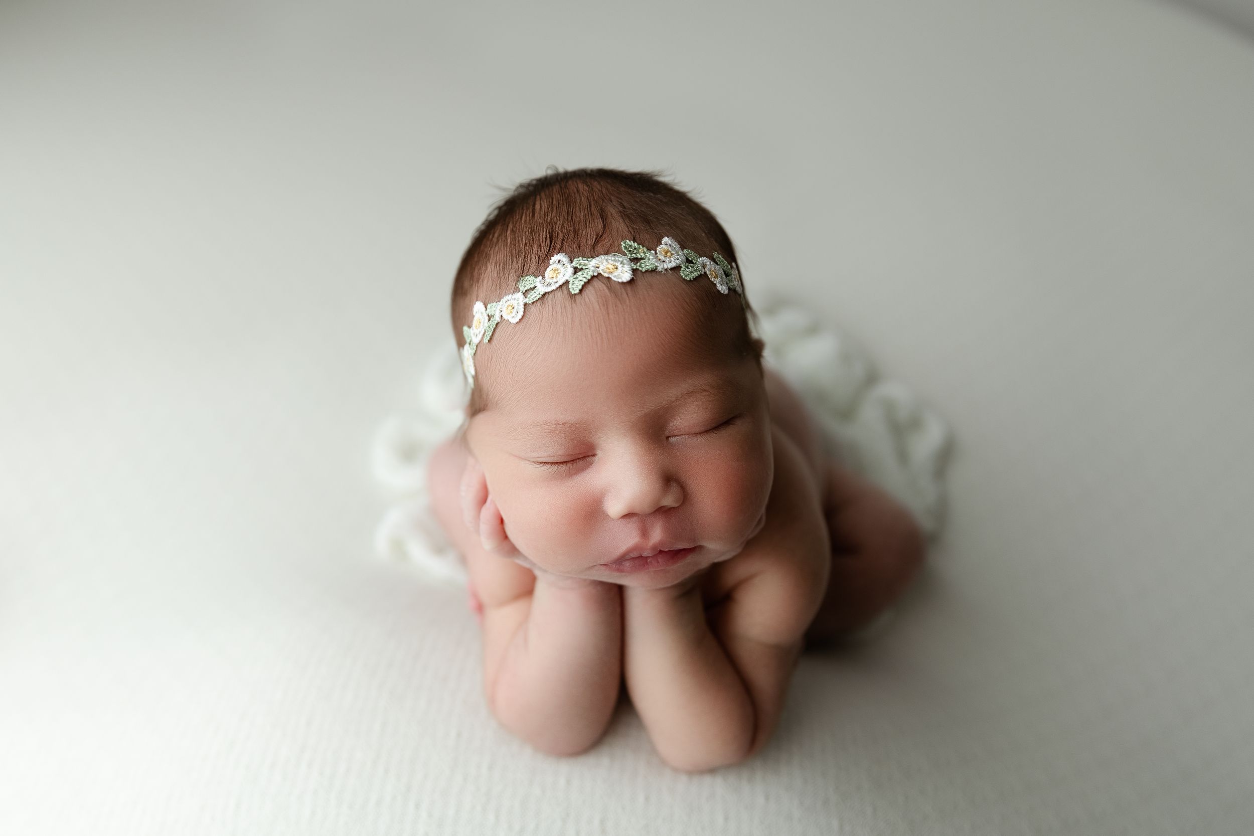 Sleeping newborn wearing delicate silver headband poses on white blanket with hands under chin.