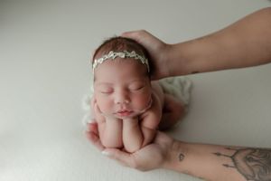 Sleeping newborn baby in fetal position wearing delicate white headband rests on parent's tattooed arms.