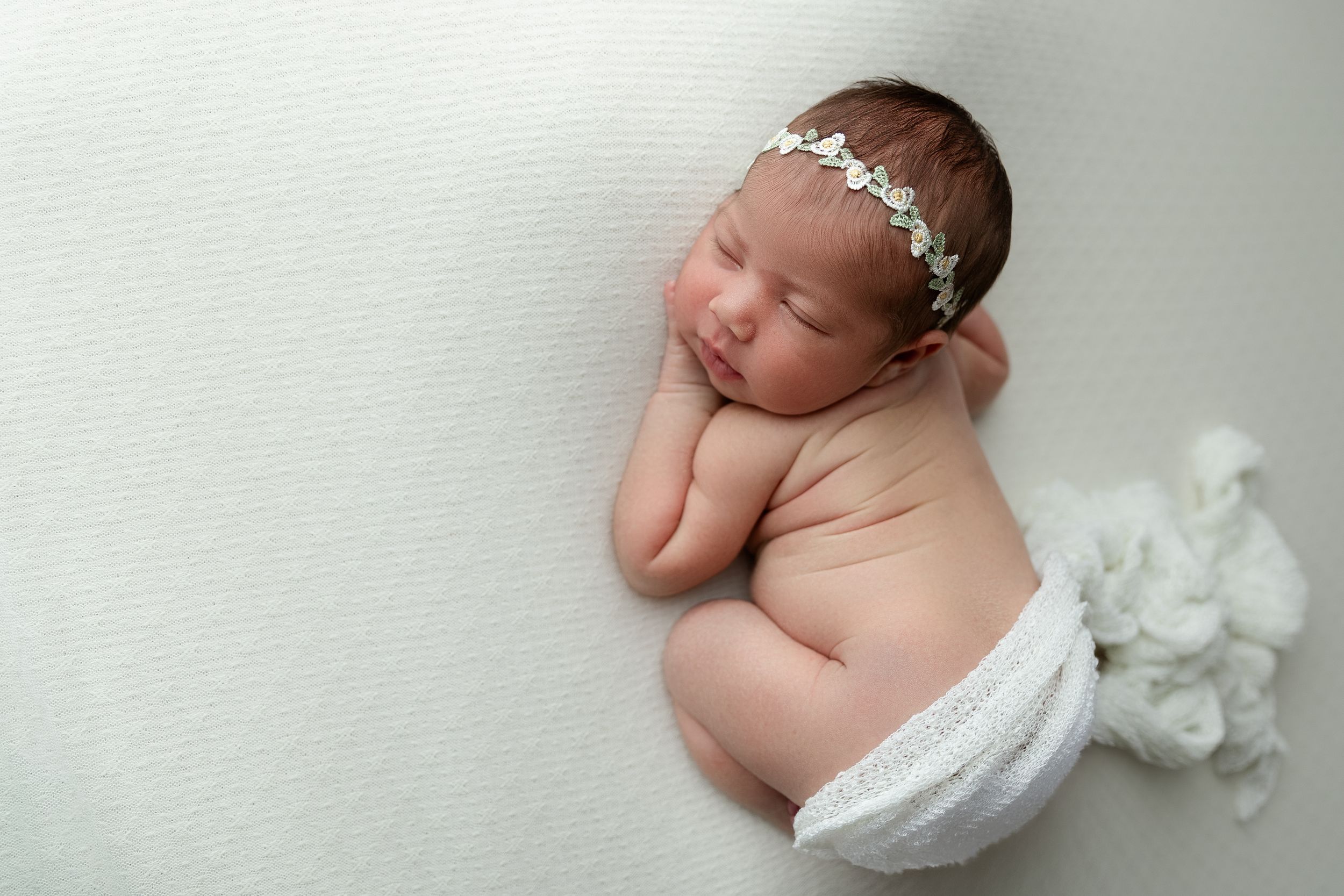 A sleeping newborn wearing a delicate white headband rests peacefully against a light backdrop with white flowers.