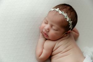 Sleeping newborn on white blanket wearing delicate floral headband in peaceful studio portrait.