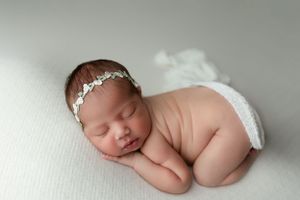 Sleeping newborn wearing delicate white headband poses on soft white blanket while curled up in peaceful pose.