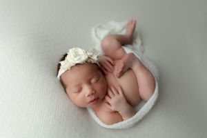 A sleeping newborn lies curled up on a soft white blanket wearing a delicate lace headband.