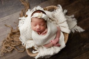 A sleeping newborn wrapped in soft white blankets and lace headband rests peacefully in a wooden basket.