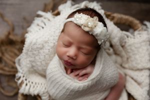 Peaceful newborn sleeping while wrapped in soft white knitted blanket with delicate white floral headband.