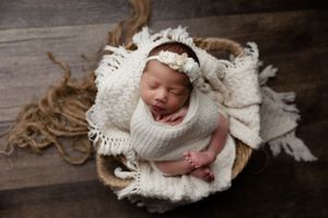 Newborn baby sleeps peacefully wrapped in white knit blanket in rustic wooden basket with textured background.
