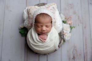 Peaceful sleeping newborn wrapped in white knitted wrap with floral bonnet on wooden floor.