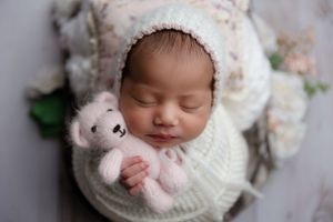 Sleeping newborn swaddled in white knit wrap holding tiny teddy bear with flowers in background.