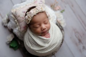 Sleeping baby wearing cream lace bonnet swaddled in white knit wrap on light wood backdrop.