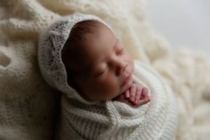 A sleeping baby wrapped in a cozy white knit blanket and bonnet lays peacefully on a cream textured backdrop.