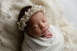 Sweet newborn baby sleeps peacefully in white knit wrap and lace crown on cream textured backdrop.