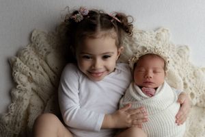 Two children lying on cream colored blanket with small one wrapped in white swaddle and wearing white long sleeve shirt.