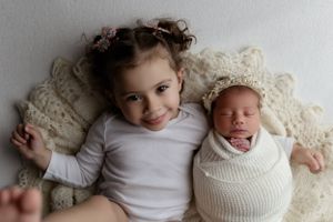 Two young children lying together on cream lace blanket with one in white knit swaddle and bonnet.