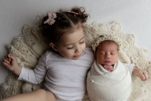 A young child and newborn baby lying together on cream-colored lace bedding in a sweet sibling portrait.