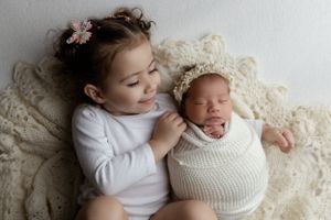 Two children lying on cream lace fabric, with one swaddled in a white knit wrap and the other wearing white long sleeves.