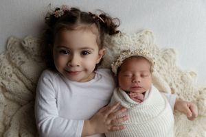 Two children resting on ivory lace, with an older sibling in white next to a swaddled newborn.