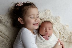 Child in white shirt lies next to swaddled newborn baby on cream lace blanket.