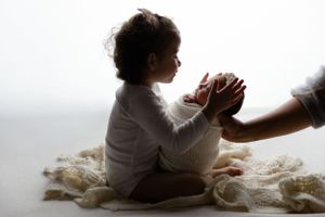 Silhouette of a young child sitting on white fabric while reaching to hold adult hands in soft lighting.