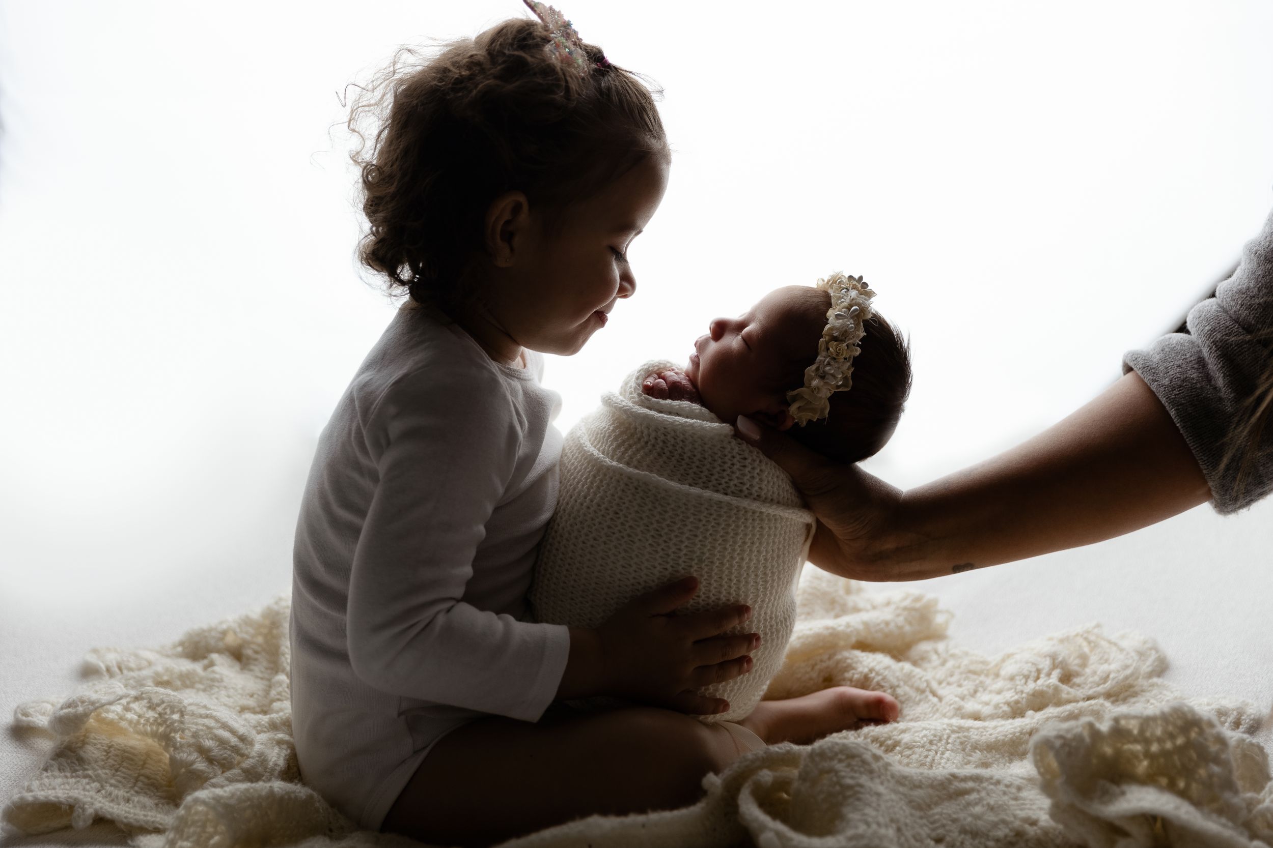 A silhouette of a tender moment shared between a child and baby wrapped in white textiles against a light backdrop.