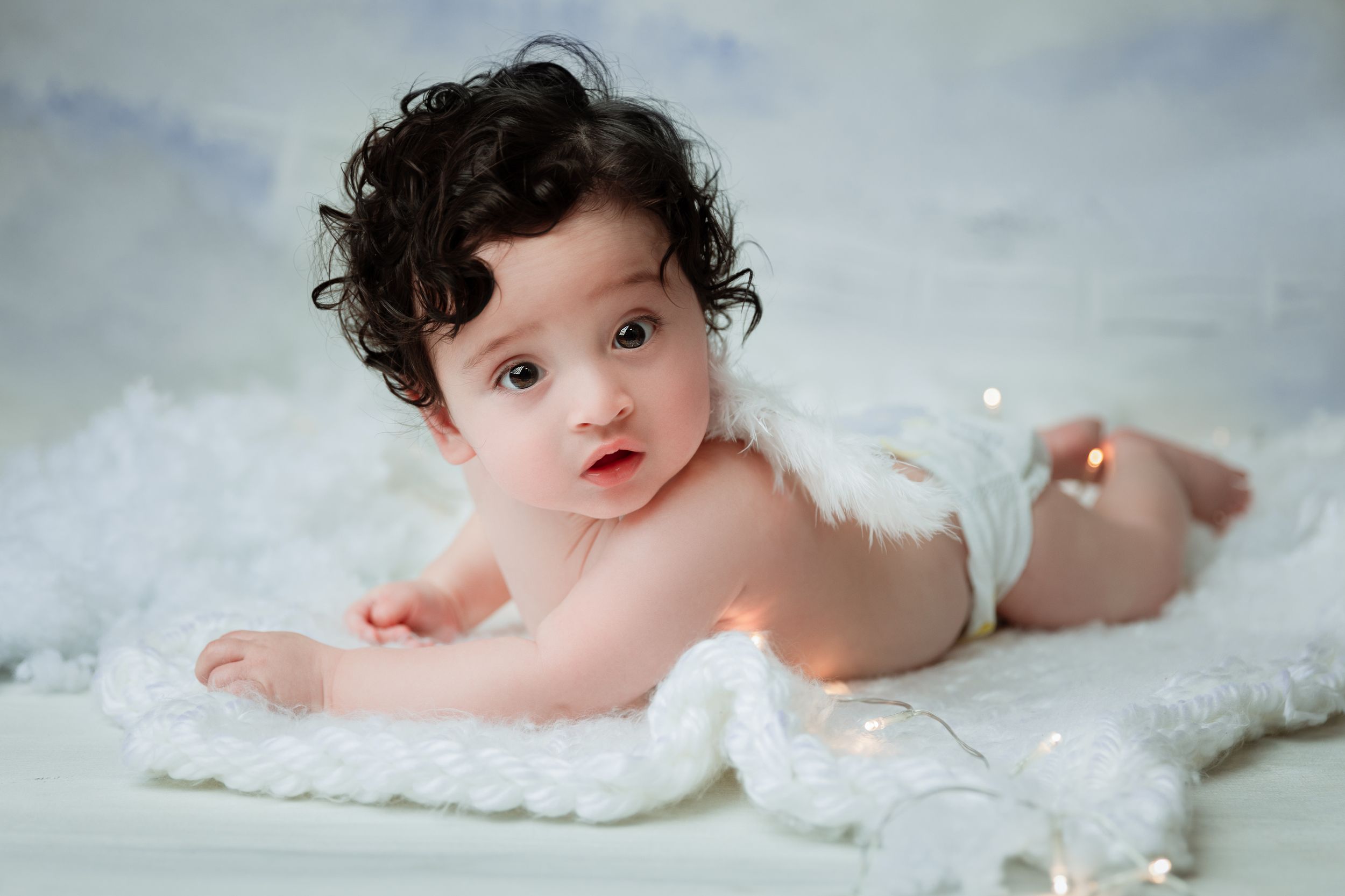 Baby with dark curly hair lying on soft white blanket wearing angel wings in gentle studio portrait.