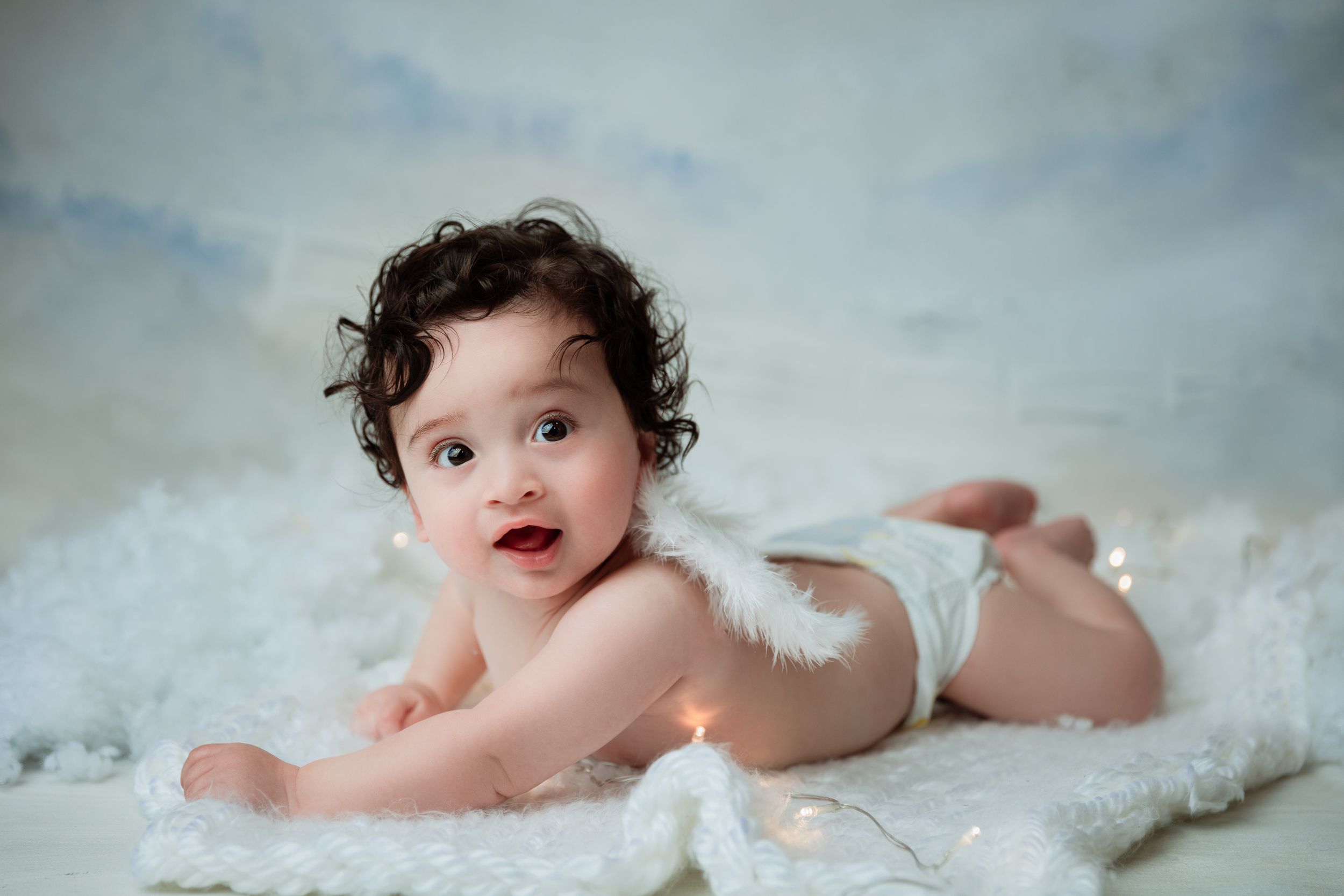 Baby wearing white angel wings lies on soft white fabric against blue background.