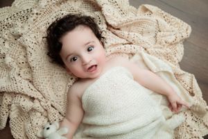 A small baby wrapped in white fabric laying on a decorative cream knit blanket.