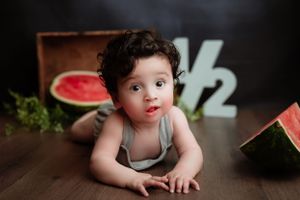 Baby in gray tank top lying on wooden floor with watermelon and decorative number 1/2 sign in background.